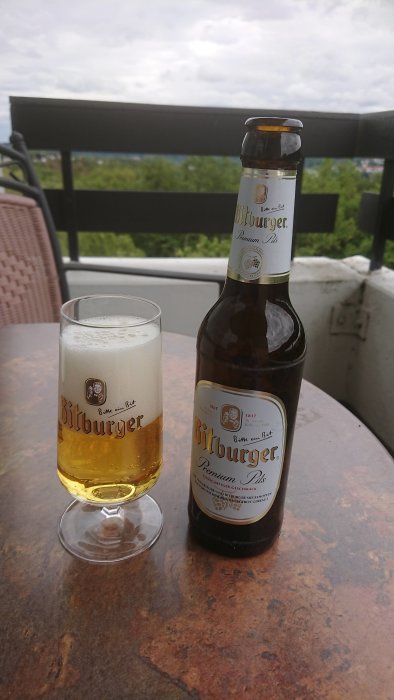 En öppen flaska av Bitburger öl och ett fullt glas på ett rostigt bord utomhus med trädtoppar i bakgrunden.