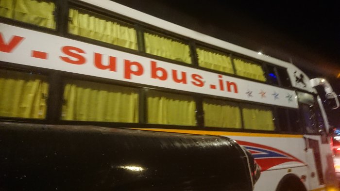 Bild på sidan av en buss märkt "V-supbus.in" med stjärnor, tagen på natten med stadsljus i bakgrunden.