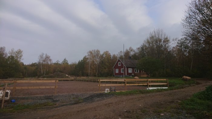 Röd stuga med vita knutar bakom en nybyggd paddock i lantlig miljö, makadamväg och staket under konstruktion.