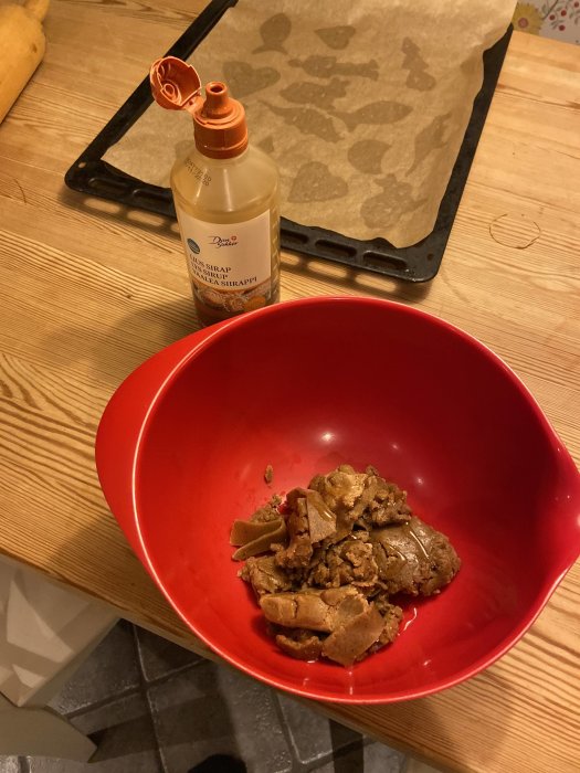 En röd skål med smulad pepparkaksdeg och en öppen sirapsflaska på ett köksbord med ett bakplåtspapper i bakgrunden.