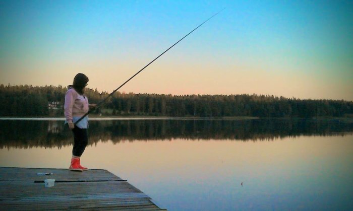 Person fiskar från brygga vid skogsbeklädd sjö i skymningen.