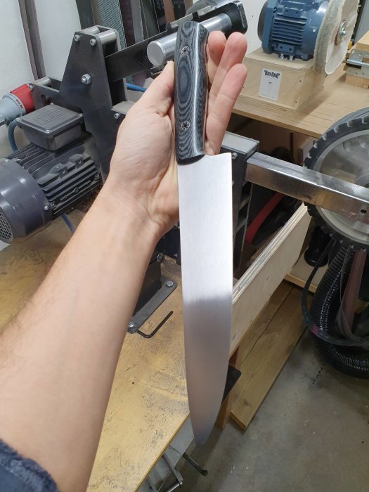 Hand håller hemgjord kockkniv med svart linnemicartahandtag i verkstad.