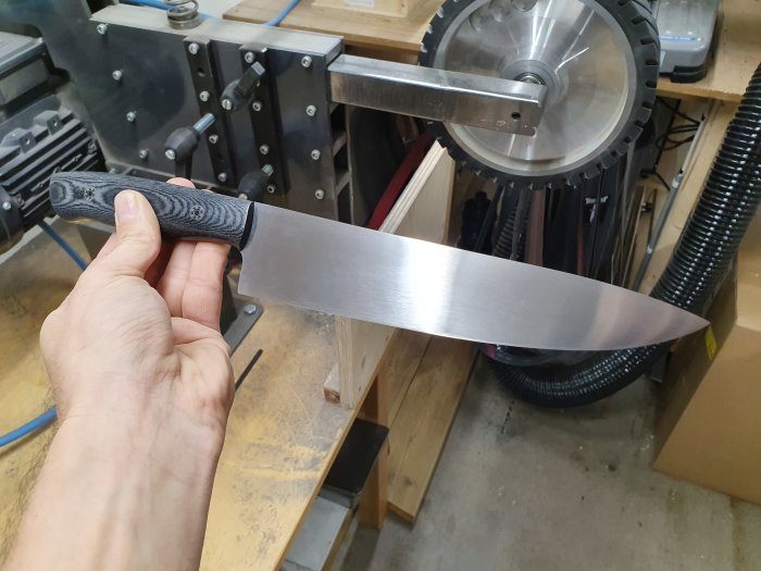 Hand håller nytillverkad kockkniv med blankt blad och svart linnemicartahandtag i en verkstad.