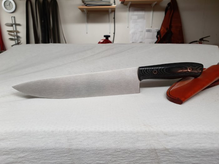 Handgjord kockkniv med silverblad och svart linnemicartahandtag, bredvid ett brunt knivfodral.