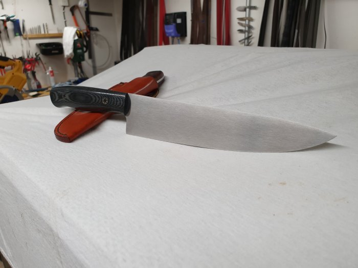 Handgjord kockkniv med RWL-34 blad och svart linnemicarta-handtag på vit bakgrund.