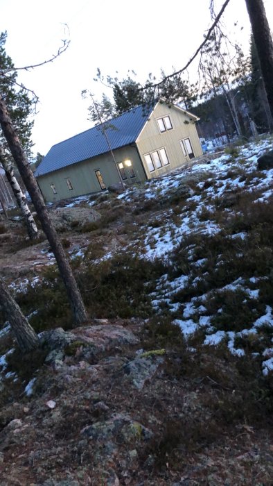 Hus med järnvitriolbehandlad fasad och svarta fönsterramar omgivet av skogsmark med lite snö.