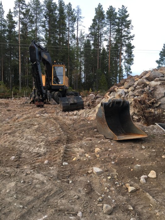 En grävmaskin på en skogstomt med uppriven mark och en hop grus och sten i bakgrunden.