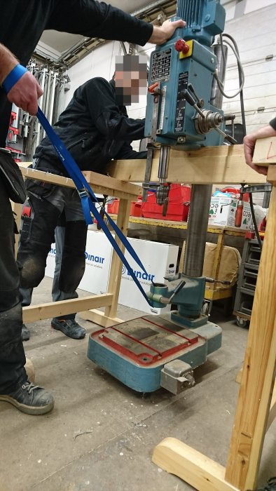 Pelarborr lyfts upp på en bjälke över bockar, med person vars ansikte är pixlat och spännband för säkerhet.