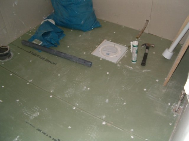 Uppbyggnadsfas av ett badrumsgolv med fuktskydd, rör och verktyg synliga på golvet.