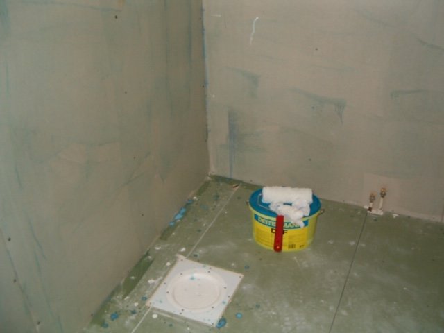 Ofärdigt badrum med gipsskivor på väggar, golvvärme och en hink med spackel.
