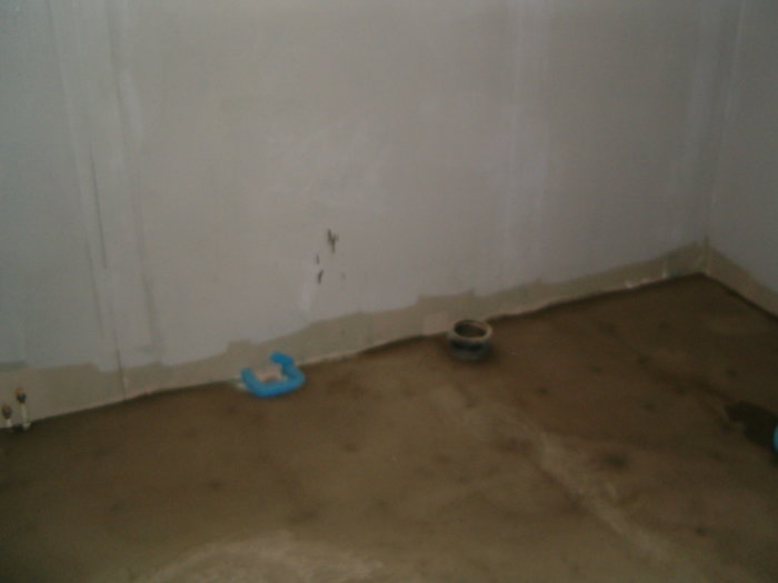 Hörn av ett rum under renovering med oskyddad golvbrunn och delvis målade väggar.