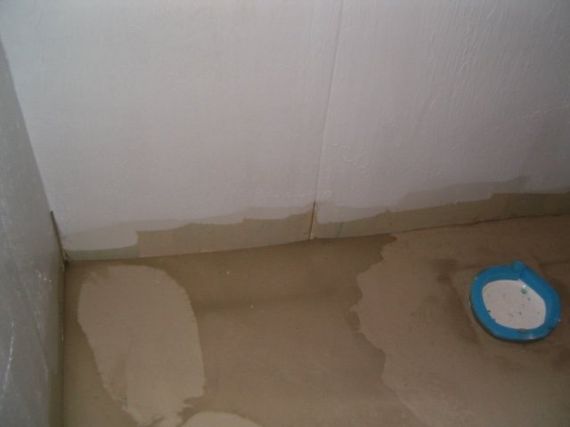 Hörn i ett rum under renovering med delvis spacklade väggar och en spackelhink på betonggolvet.