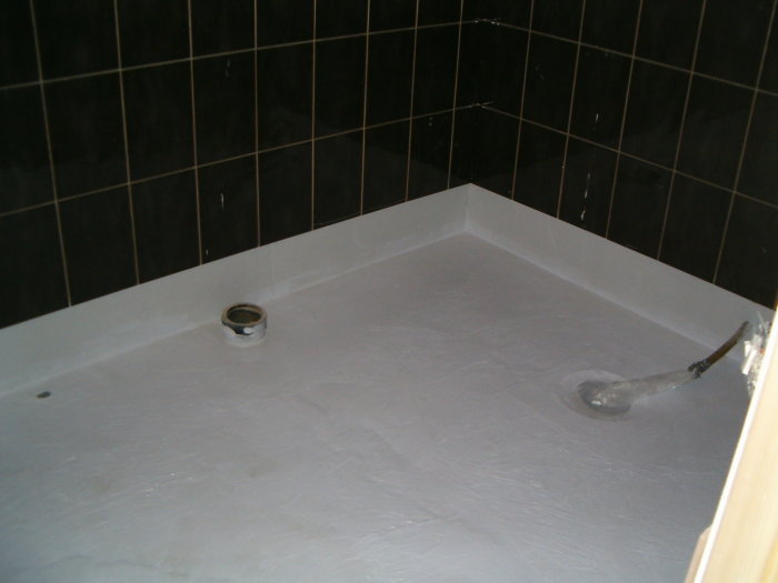 Nyinstallerat duschgolv i betong med avlopp och svart kakel på väggarna.