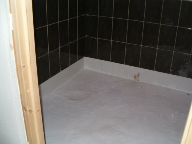 Halvfärdigt badrum med vita golv och svarta väggkakel, synlig VVS-installation, utan sanitetsgods.