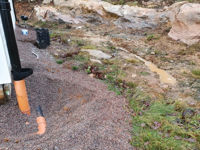 Stående vatten nära husets grund med synlig avloppsrör och en sluttning med berg i bakgrunden.