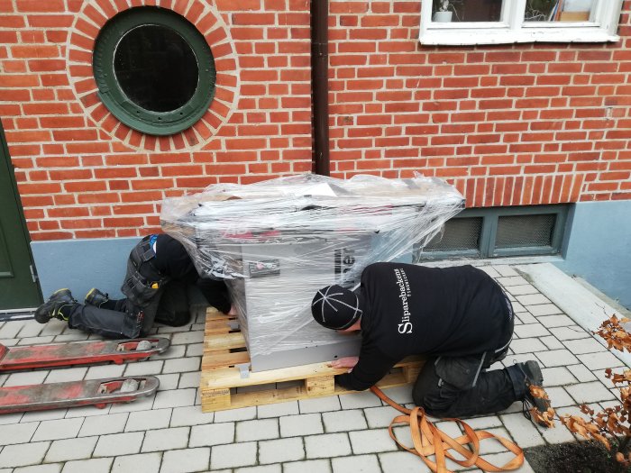 Två personer installerar tung maskin på pall med hjälp av spännband och verktyg utanför byggnad.