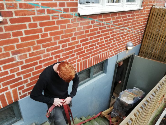 Person justerar spännband vid lastning av gods i källaringång, kollega övervakar nedanför.