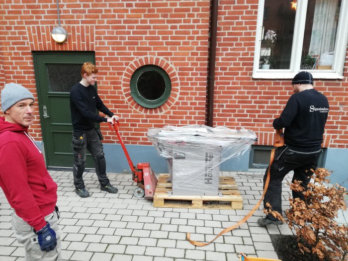 Arbetsteam använder pallbock för att flytta ett maskineripaket framför en tegelbyggnad.
