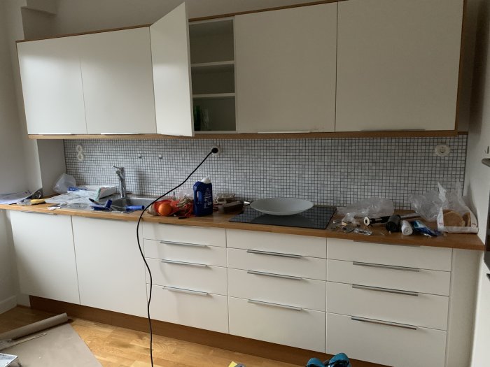 Köksinredning med vit flagnande kökslucka ovanför spisen, små kakelplattor som stänkskydd och rörigt köksbänk med verktyg.