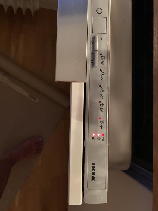 Diskmaskinslucka från IKEA med kontrollpanel och symboler, inklusive en fot i bakgrunden.