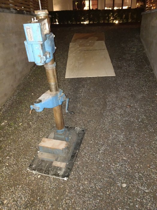 En begagnad blå Arboga pelarborr står på en plattform med hjul på en grusig yta med plywoodbanor framför.