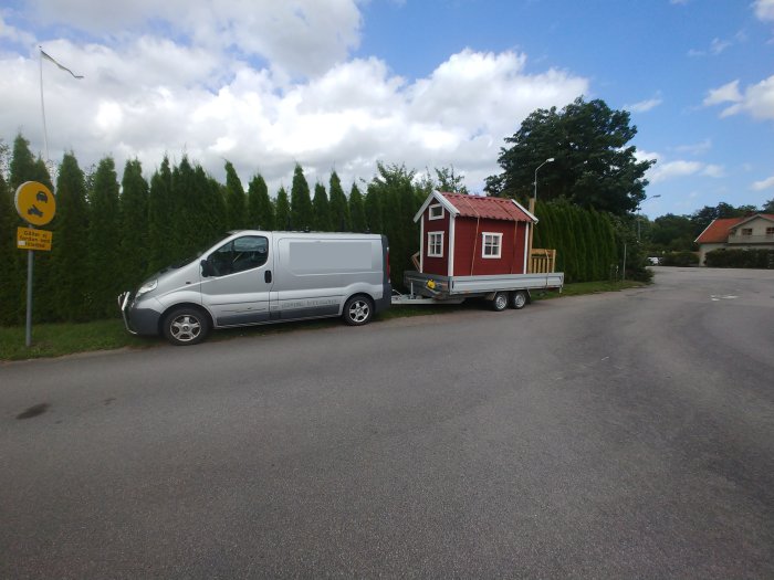 Skåpbil drar en släpvagn med en röd lekstuga längs en gata.