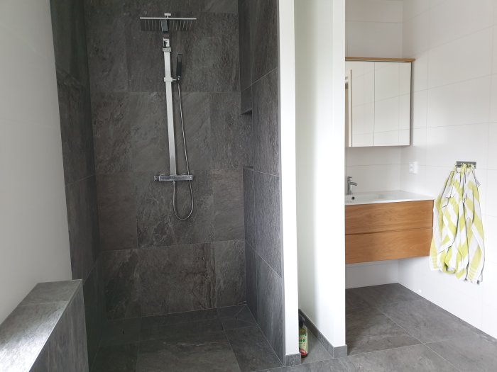 Modernt badrum med gråa kakelväggar, duschhörna och träfärgat badrumsskåp.