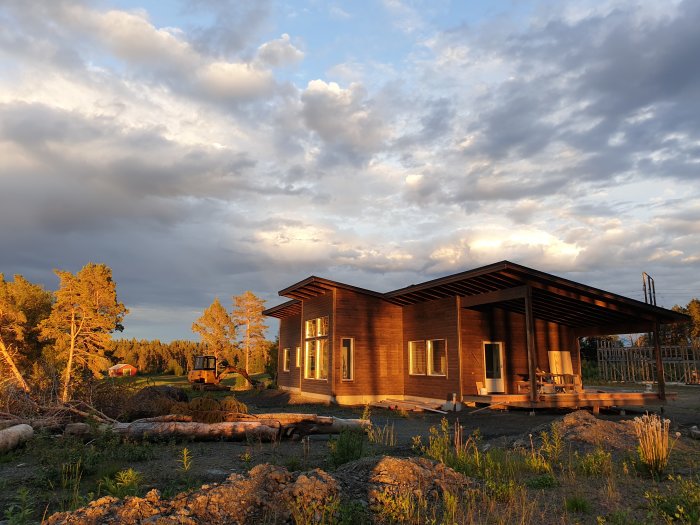 Nybyggt trähus i solnedgång med byggmaterial och en grävmaskin i bakgrunden.