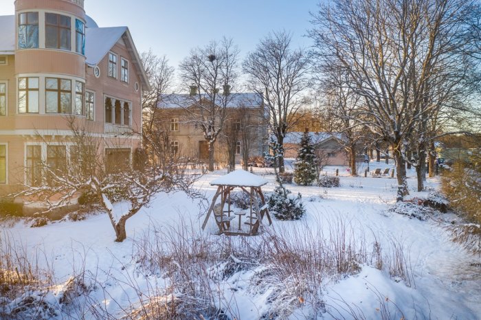 Vinterträdgård med villa, snötäckt mark och vedergälla i solljus.