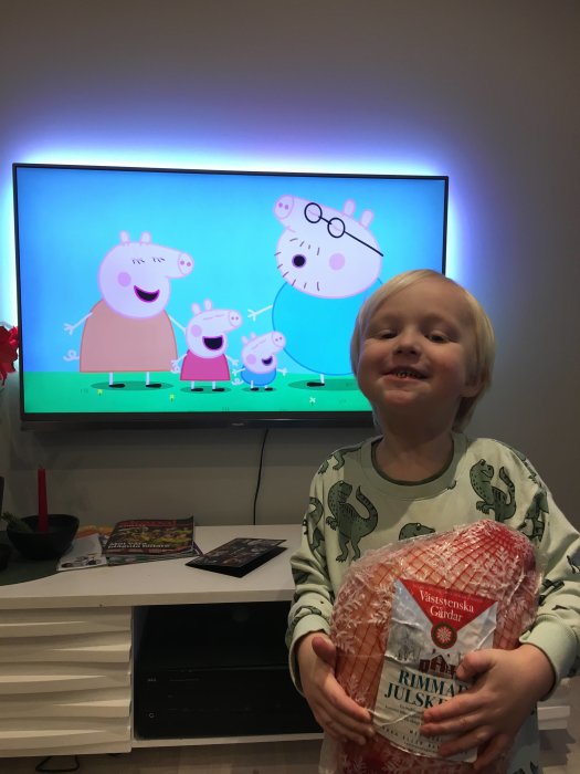 Leende barn håller i en skinka framför TV:n som visar Peppa Pig under en julbelysning.