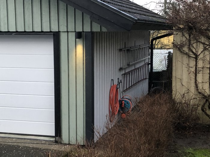 Garage med upphängda slangvindor och stegar på sidoväggen, omgivet av grönska.