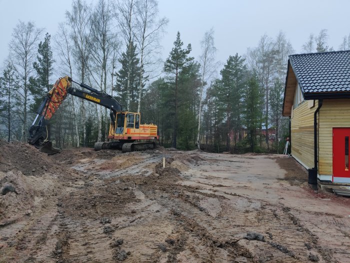 Grävmaskin som arbetar med markarbeten nära ett gult hus i dagsljus.