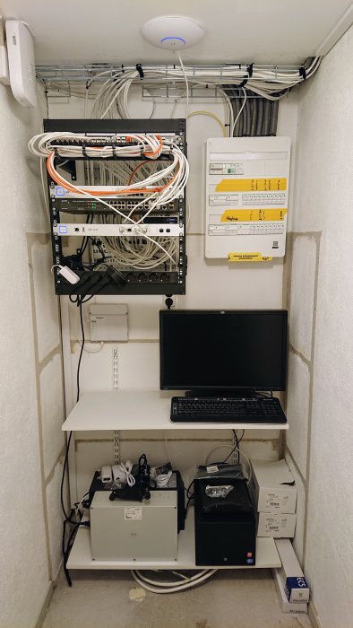 Teknikrum med flera patchpaneler, nätverksutrustning, en accesspunkt i taket och en dator på ett skrivbord.