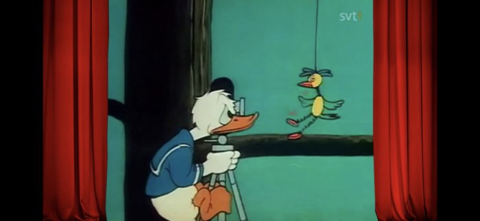 Animerad figur av en vit anka med sjömanskostym använder ett kamerastativ, står på en scen bredvid en fågel.