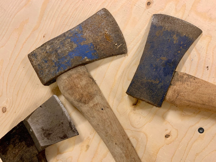 Tre använt utseende yxor med olika grad av slitage och rost ligger på en ljus träyta.