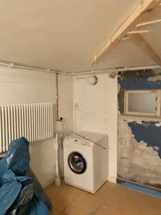 Källartvättstuga med vit tvättmaskin, sliten vägg med flagnande färg och ojämn yta, synlig rörledning och radiator.