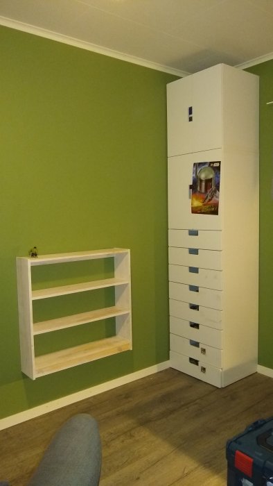 Ny inredning i sonens rum med ett högt vitt skåp och en tom hylla mot en grön vägg.