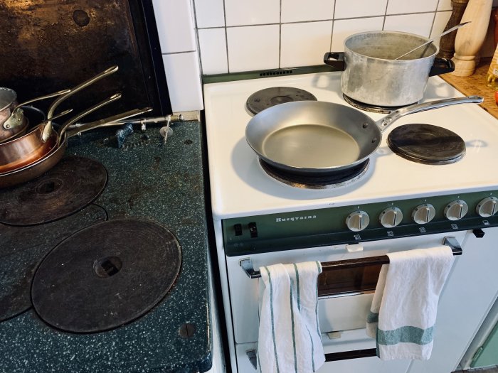 En ny de Buyer stålpanna på en Husqvarna spis bredvid andra köksredskap och en gammal kastrull.