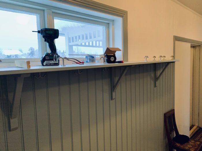 Kapprum med nyinstallerade ankarkrokar under ett fönster, bredvid en bordsyta med en skruvdragare.