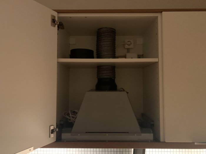 Installationsvy av en köksfläkt, rör och uppfälld plåt underifrån i ett köksskåp.