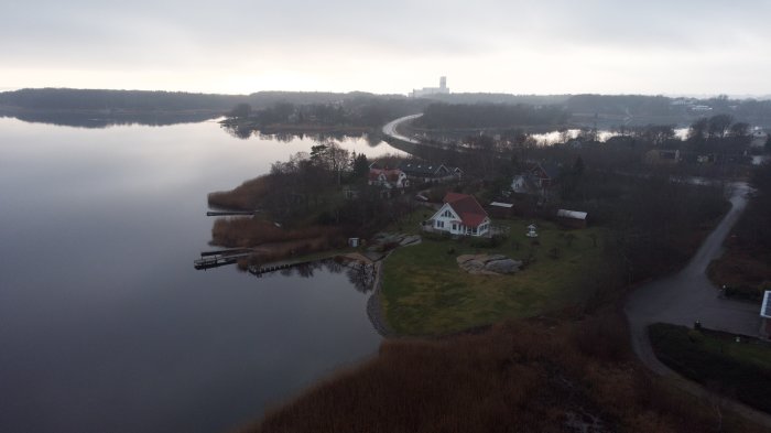 Drönarbild på ett sjönära bostadsområde med hus och vägar vid skymningen.