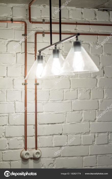 Modern belysning med tre triangelformade lampskärmar hängande från svarta kablar framför en vit tegelvägg med synlig kopparfärgad rörinstallation.