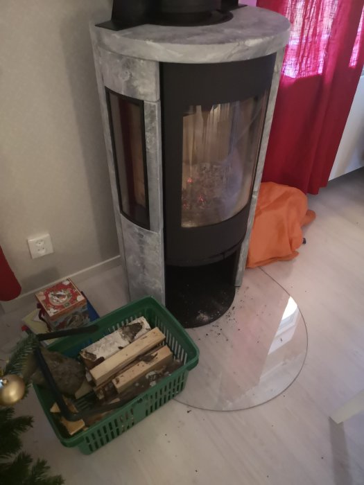 Modernt kakelugn med ved i grön plastkorg och aska på golvet, nära julgran.