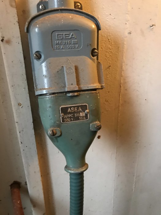 Vintage ASEA elkomponent med en grön, flexibel slang fäst på en vit vägg, eventuellt till en kompressor från 70-talet.