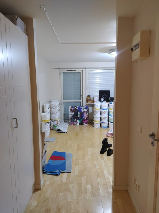 Lägenhetsrum använd som ateljé med byggmaterial och hinkar staplade längs väggen, oplastad yta före renovering.
