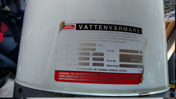Upp och ner bild av en vattenvärmare med tekniska specifikationer och en varningsetikett synlig.