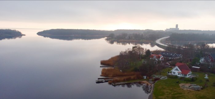 Drönarvy över sjö vid gryning med spegelblankt vatten, bostadshus och en väg i landskapet.