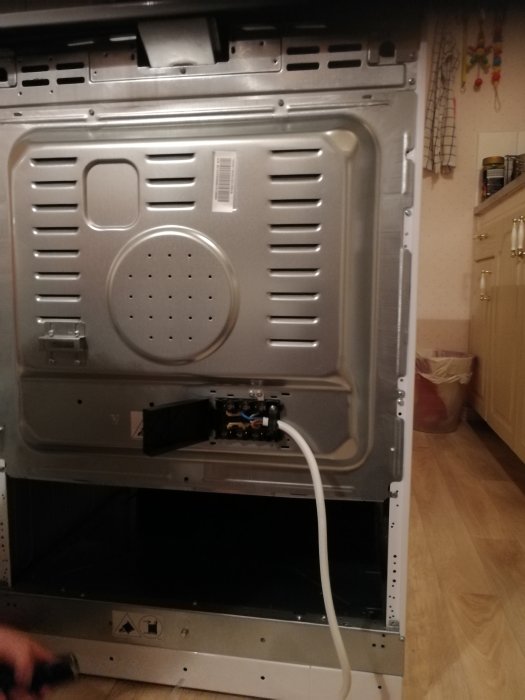 Baksidan av en ugn med en vit elkabel ansluten, i ett kök med synligt vägguttag och kökstillbehör.