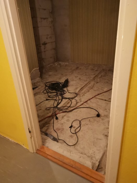 Rum under renovering med betonggolv och vita väggar, synliga träreglar och strömkablar på golvet.