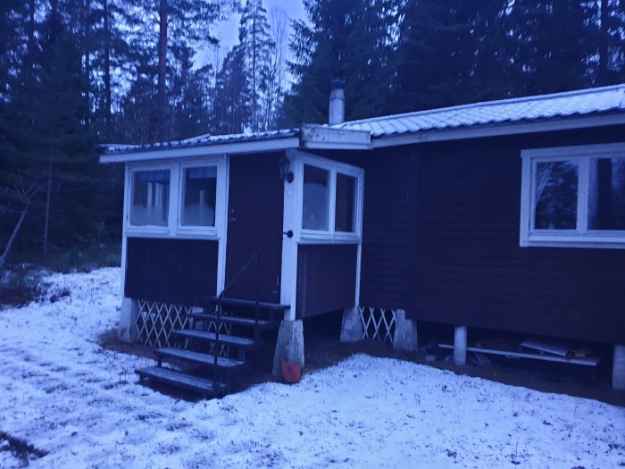 Delvis snötäckt mark framför ett mörkfärgat hus med vit inbyggd veranda och utrymme under huset.